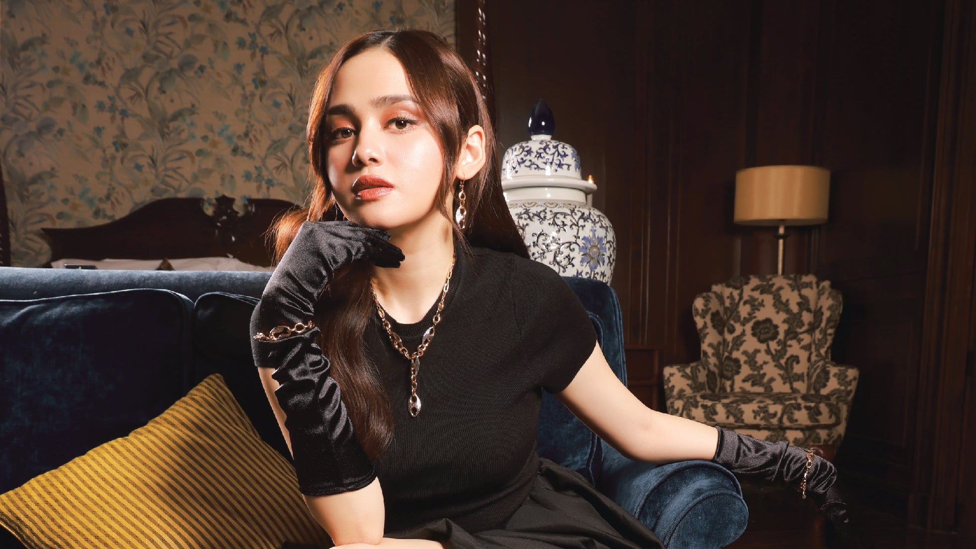 Woman in a black dress and knee-high boots sitting on a blue velvet couch in an elegant room.